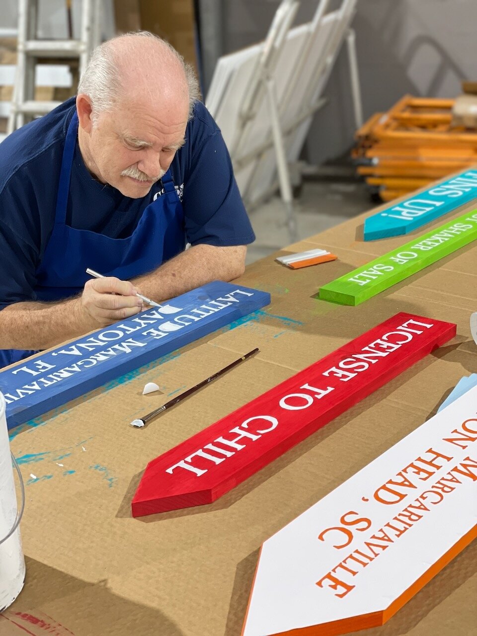 CG Signs team member hand-painting Latitude Margaritaville carved signs — License to Chill, Daytona, Hilton Head — in the Orange Park shop