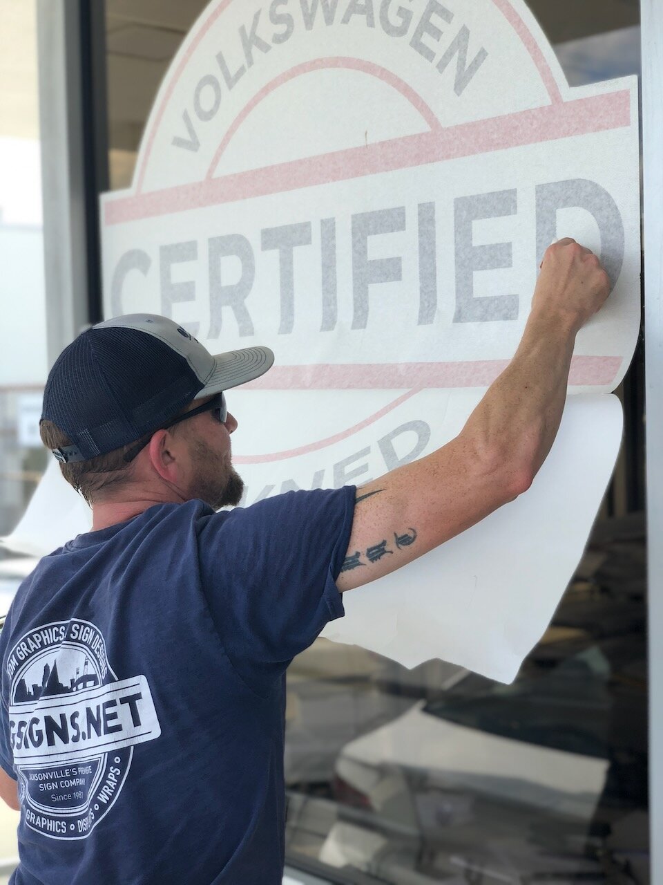 CG Signs installer applying a Volkswagen Certified vinyl graphic to a dealership window, wearing branded company shirt